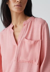Blusa rosa con texture, caratterizzata da un colletto alla coreana, una parte di abbottonatura e una tasca sul petto. Il tessuto ha un aspetto morbido e leggero.