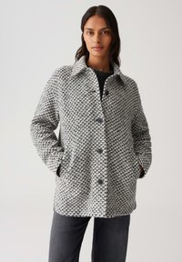 Frau trägt einen geknöpften, grau-weißen strukturierten Wollmantel mit Händen in den Taschen, kombiniert mit dunkelgrauen Hosen vor einem schlichten Hintergrund.