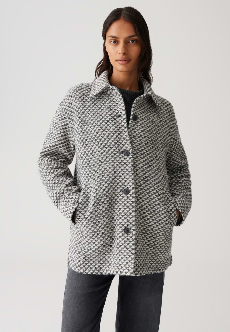 Frau trägt einen geknöpften, grau-weißen strukturierten Wollmantel mit Händen in den Taschen, kombiniert mit dunkelgrauen Hosen vor einem schlichten Hintergrund.