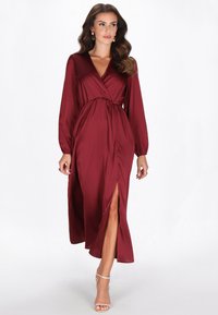Bordowa sukienka kopertowa z jedwabistej tkaniny, z długimi rękawami, głębokim dekoltem w serek, marszczonym pasem i bocznym rozcięciem, zestawiona z sandałkami na cienkich paskach.
