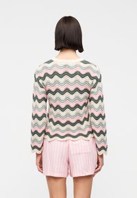 Γυναί κα με ίσια σκουρόχρωμα μαλλιά που φοράει ένα πλεκτό πουλόβερ με κυματιστές ρίγες σε πολλά χρώματα και ανοιχτό ροζ ριγέ σορτς, εμφανιζόμενη από πίσω.