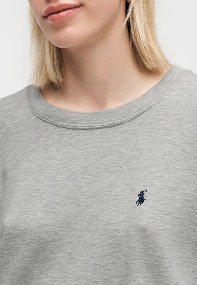 Γυναίκα που φοράει ένα ανοιχτό γκρι πουκάμισο με λαιμόκοψη, με ένα μικρό σκοτεινό μπλε λογότυπο παίκτη πόλο στο στήθος, εμφανιζόμενη από το λαιμό μέχρι τη μέση.