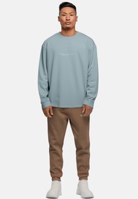 Felpa azzurra chiara con collo rotondo e logo ricamato. Abbinata a pantaloni della tuta marroni e sneakers bianche. Design semplice e sportivo.