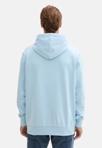 Felpa blu chiaro con un ampio cappuccio, texture morbida e polsini e orlo a coste. Stilata con una vestibilità rilassata, indossata sopra jeans scuri.