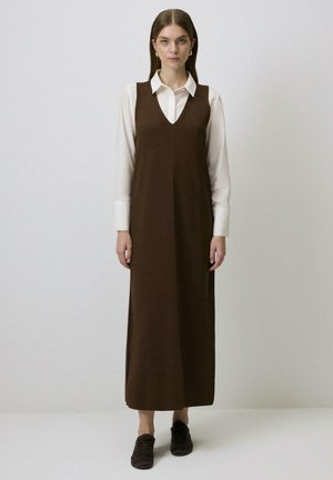Robe sans manches marron avec un col en V, atteignant les chevilles, portée par-dessus une chemise blanche. Texture lisse, silhouette décontractée, associée à des chaussures sombres.