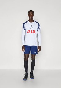 Maillot blanc à manches longues avec des accents bleus et le logo "AIA", associé à un short bleu marine et des chaussettes de football noires. Présente la marque Nike.