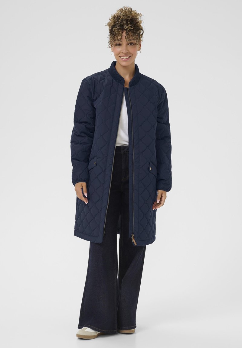 Femme aux cheveux bouclés portant un long manteau matelassé bleu marine, une chemise blanche, un pantalon large foncé et des chaussures beige, souriante et debout devant un fond gris.