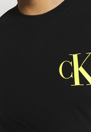 Nahaufnahme eines schwarzen Rundhalsausschnitt-Shirts mit einem gelben "cK"-Logo auf der oberen linken Brust.