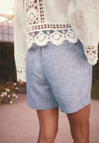 Lyseblå linnedshorts med tekstureret overflade, der har en baglomme, parret med en hvid broderet blondeoverdel med bølgede kanter.