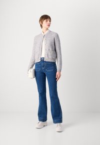 Light gray knitted cropped cardigan with gold buttons, paired with blue flared jeans. Model holds a small white bag, wearing white sneakers.