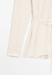 Blouse crème en tissu léger, dotée de manches longues, d'une taille ceinturée et d'un ourlet asymétrique avec une texture lisse.