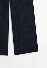 Pantalons en denim bleu foncé, jambes posées à plat sur une surface blanche, montrant l'ourlet inférieur et la coupe droite.