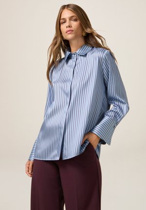 Camicia azzurro chiaro con strisce verticali bordeaux, colletto classico e maniche larghe, abbinata a pantaloni a gamba larga bordeaux scuro.