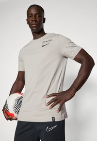 Camiseta de algodón gris con cuello redondo, que presenta los logotipos negros de "Spurs" y Nike. Modelo sosteniendo un balón de fútbol en blanco y negro, vistiendo pantalones oscuros.