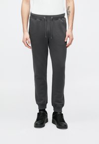 Szürke pamut melegítőnadrág elasztikus derékpánttal és húzózsinórral, bordázott mandzsettákkal és oldalsó varrásokkal. Fekete tornacipővel párosítva.