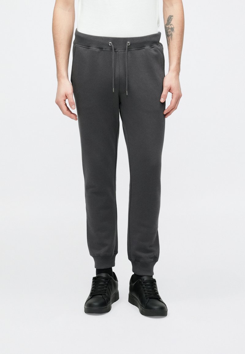 Szürke pamut melegítőnadrág elasztikus derékpánttal és húzózsinórral, bordázott mandzsettákkal és oldalsó varrásokkal. Fekete tornacipővel párosítva.