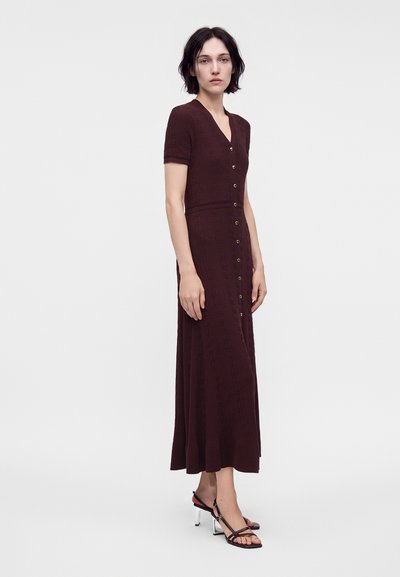 Femme debout portant une robe marron foncé à manches courtes boutonnée et des sandales à talons hauts noires avec des talons blocs transparents sur fond blanc.
