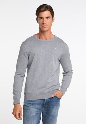Mann mit kurzen braunen Haaren, der einen grauen Langarmpullover und blaue Jeans trägt, steht mit einer Hand in der Tasche vor weißem Hintergrund.