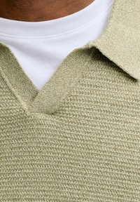 Pull-over vert clair en maille avec un col en V et une surface texturée, superposé sur un t-shirt blanc à col rond. Col et ourlet côtelés.