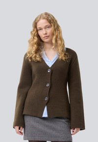 Ung kvinde med langt krøllet blond hår iført en brun knapkort cardigan, lyseblå skjorte og mønstrede nederdel mod en ensfarvet baggrund.