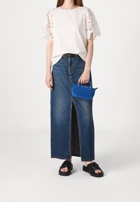 Biała koszulka z koronkowymi rękawami, jeansowa spódnica o średnim stanie z przednim rozcięciem oraz czarne sandały na platformie. Trzyma małą niebieską torebkę.