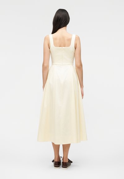 Γυναίκα με μακριά σκουρόχρωμα μαλλιά, που φορά μπλούζα χωρίς μανίκια σε εκρού χρώμα και καφέ δερμάτινα παπούτσια με κορδόνια, στέκεται γυρισμένη προς τα πίσω μπροστά σε μονόχρωμο φόντο.