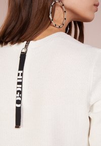 Donna con capelli castani che indossa grandi orecchini a cerchio in argento e un maglione bianco con texture e una cerniera nera sul retro.