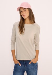 Young woman wearing a light pink cap, beige long-sleeve shirt, and blue jeans with hands in pockets, against a plain light background.