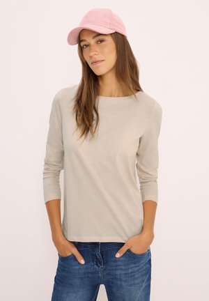 Jeune femme portant une casquette rose clair, une chemise à manches longues beige et un jean bleu, les mains dans les poches, devant un fond clair.