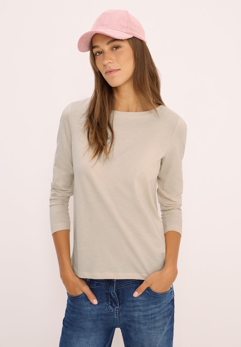 Young woman wearing a light pink cap, beige long-sleeve shirt, and blue jeans with hands in pockets, against a plain light background.