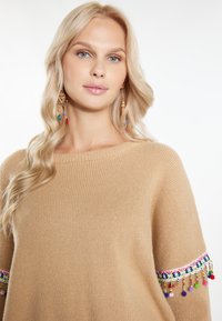 Beige gebreide trui met een ruime halslijn en kleurrijke geborduurde accenten op de mouwen, met kleine decoratieve franjes.