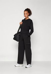 Sieviete ar lokainu astīti, ģērbta melnā rāvējslēdzēja džaketē un platajās biksēs, baltās sporta kurpēs, nes melnu pleca somu, stāvot uz viendabīgas fona.