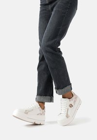 Sneakers platform bianchi con un design testurizzato, arricchiti da un accento metallico e dettagli beige chiaro, abbinati a jeans scuri tagliati.
