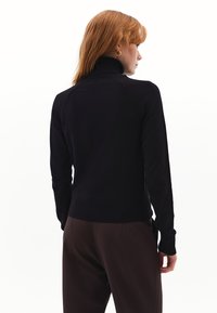 Schwarzer Pullover mit hohem Kragen und geripptem Saum sowie Bündchen, der eine glatte Textur und eine figurbetonte Passform aufweist. Trägt braune Hosen und wird von hinten betrachtet.