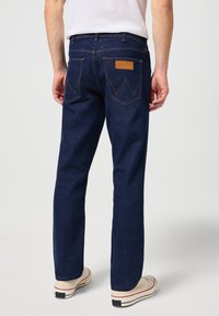 Donkerblauwe spijkerbroek met rechte pijpen, voorzien van een leren label, gedefinieerde achterzakken met een duidelijk patroon en contrasterende stiksels.