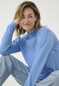 Ljusblå stickad tröja med långa ärmar och hög krage, med ribbade muddar och avslappnad passform, i kombination med ljusa denimjeans.