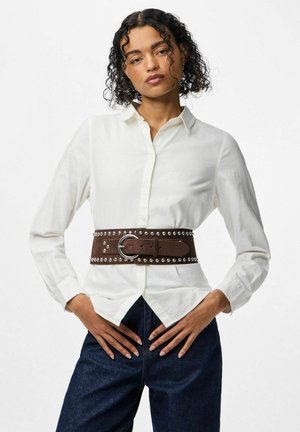 Une ceinture en cuir brun avec des clous en argent et une boucle ronde, portée par-dessus une chemise blanche à boutons, au-dessus d'un pantalon en jean foncé.