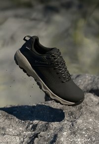 Schwarze Wanderschuhe mit Mesh-Einsätzen, strapazierfähiger Gummisohle und grauen Akzenten. Verfügt über ein Schnürdesign und ein Logo an der Seite. Über einem felsigen Untergrund erhöht.