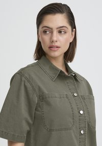 Jeune femme aux cheveux bruns courts portant une chemise olive à boutons avec deux poches sur la poitrine, regardant sur le côté devant un fond uni.