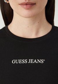 Fekete pamut póló rövid ujjakkal, kerek nyakkivágással, elöl fehér „GUESS JEANS” logóval. Simább textúrájú.