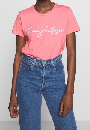 Osoba nosząca różowy t-shirt Tommy Hilfiger wpuszczony w niebieskie, wysokie jeansy, stojąca na jednolitym tle.
