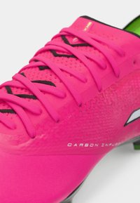 Třpytivě růžový fotbalový kopač s texturovaným svrškem, černými tkaničkami a akcenty. Na boku je viditelný nápis "Carbon Infused".