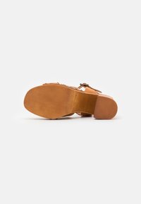 Sandale avec semelle lisse couleur tan et talon bas, montrant une lanière en cuir avec boucle, présentée sur fond blanc avec la semelle tournée vers le haut.
