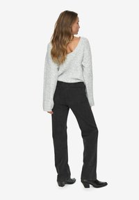 Frau mit langen Haaren, die einen hellgrauen Strickpullover mit tiefem Rückenausschnitt, schwarze Jeans und schwarze Stiefeletten trägt, seitlich stehend.