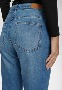 Modre hlače z visokim pasom, pet žepov, šivi na zadnjem žepu in rjava usnjena oznaka na pasu. Mehka denimska tekstura.