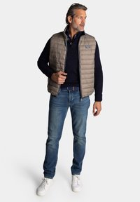 Beige gewatteerde vest met hoge kraag, gedragen over een marineblauw sweater met een gestructureerd patroon, gecombineerd met blauwe jeans en witte sneakers.