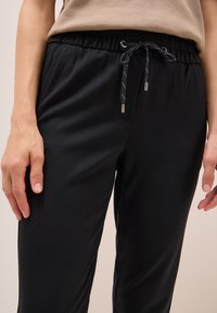 Nahaufnahme einer Person, die schwarze Jogginghosen und ein beiges Oberteil trägt, mit entspannten Händen an den Seiten vor einem neutralen Hintergrund.