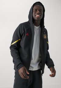Schwarze Zip-Jacke mit Kapuze, mit rotem Adidas-Logo, goldfarbenen Akzenten an den Ärmeln und einem Atlanta United-Patch auf der Brust.