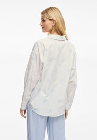 Camicia bianca con bottoni caratterizzata da modelli di ancore azzurro chiaro, maniche lunghe e colletto classico. Il tessuto appare liscio e leggermente trasparente.