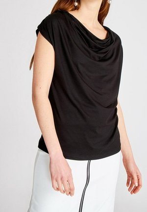 Vrouw draagt een zwarte gedrapeerde top met capuchonhals en korte mouwen en een witte rok met verticale rits aan de voorkant.